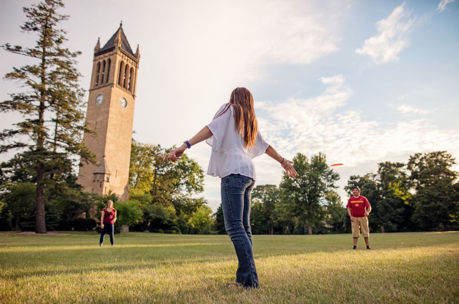 Focus on one student, facing away from the camera, throwing a frisbee to two friends in front of the campanile 