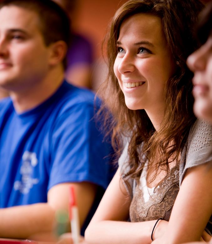 A Female student, smiling and looking to the left of the camera at in class as they are listening to lecture