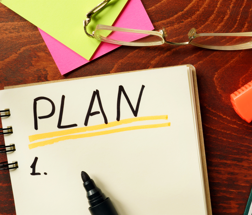 Overhead photo of a desk including a notebook with large block letters saying PLAN, highlighters, glasses, and sticky notes