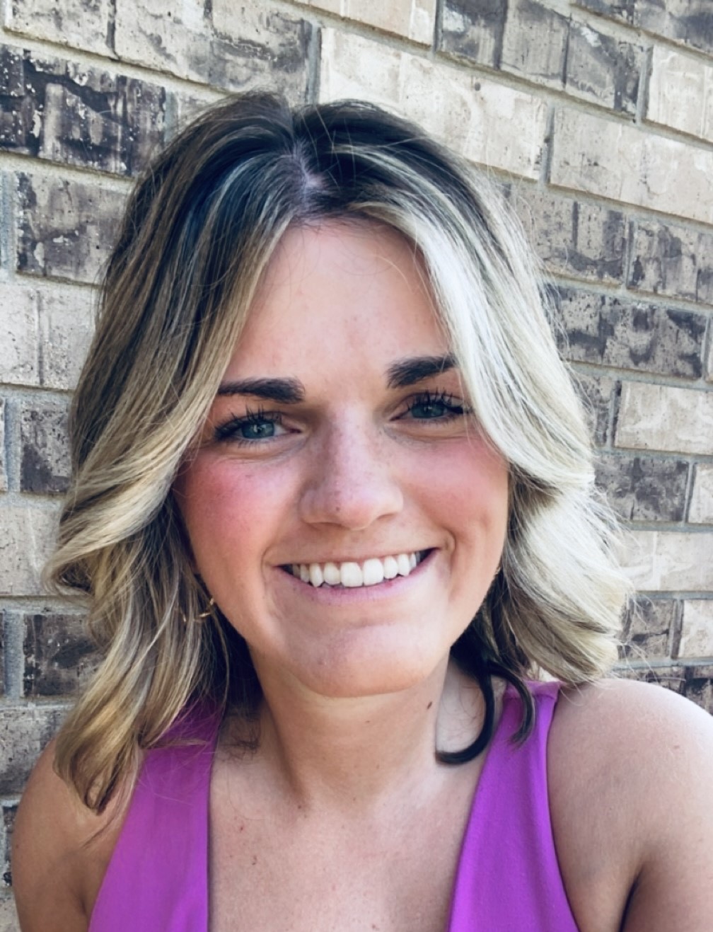 A picture of a young woman with blonde hair, wearing a pink tank top, smiling in front of a brick wall. 