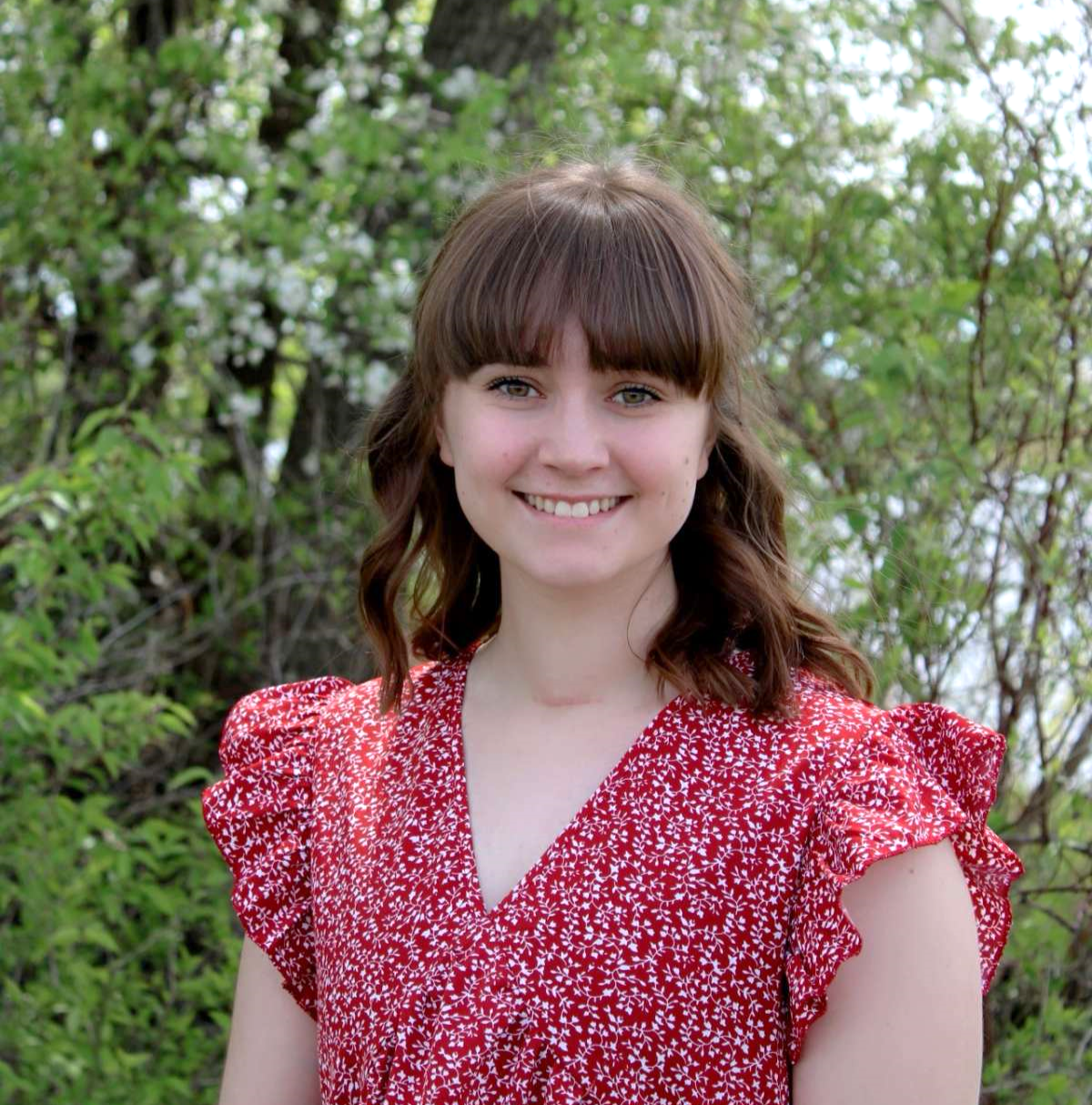 A picture of Sydney wearing a red dress in front of a green bush, smiling. 