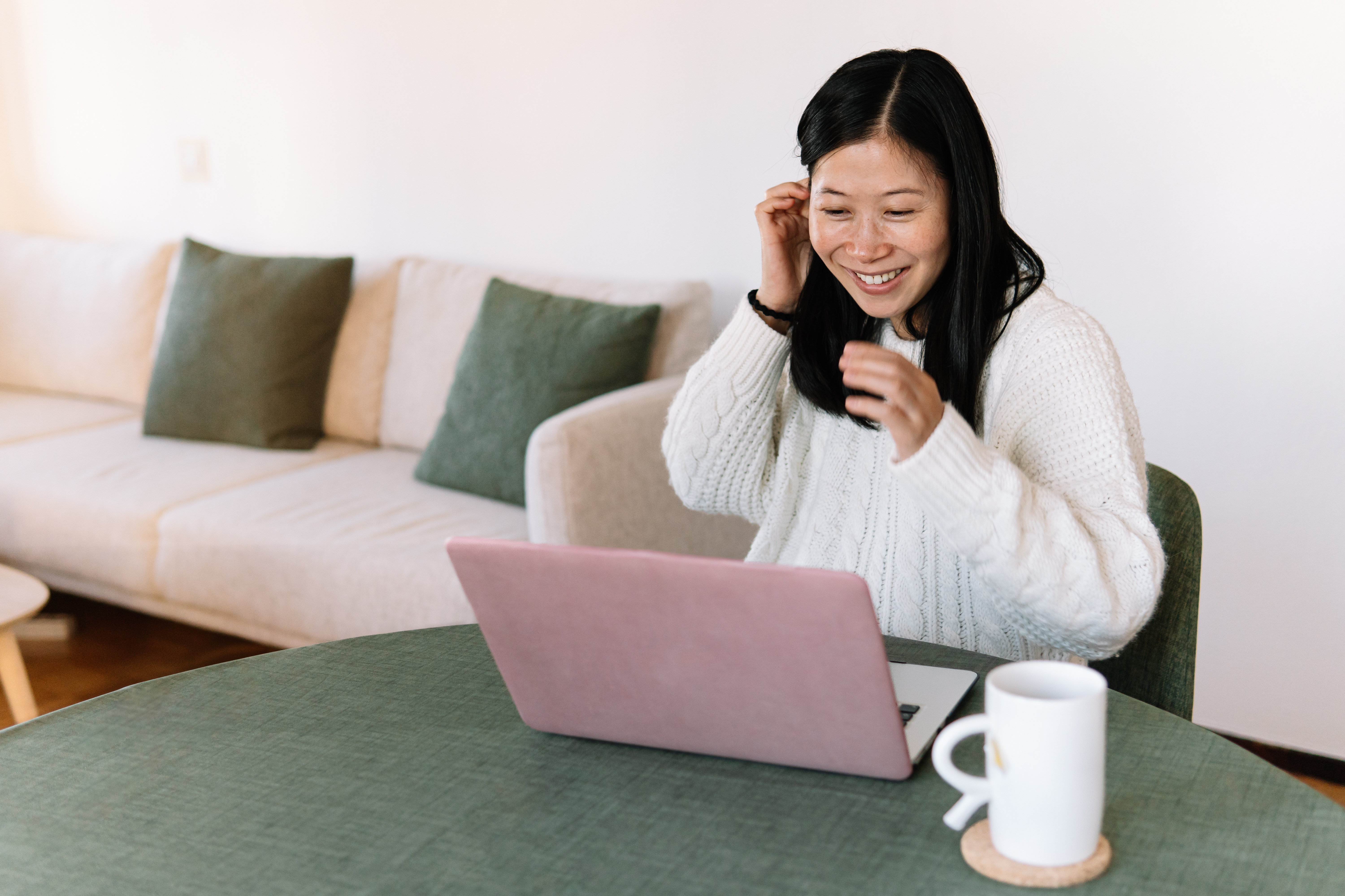 Woman, Asian with black long hair, is smiling while sitting at a table with her laptop and coffee