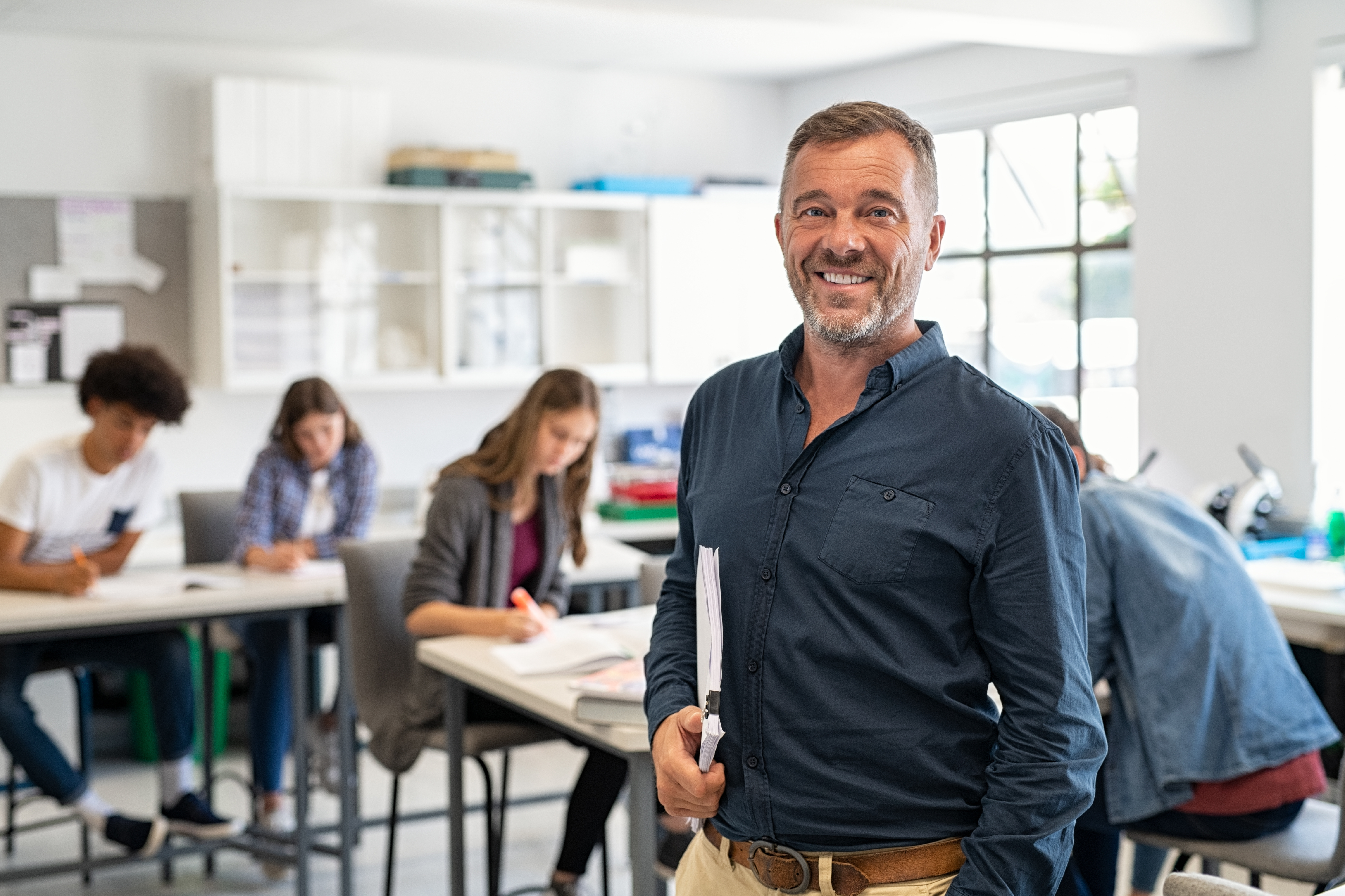 A male instructor wearing a blue button-up shit, holding a stack of papers, smiling at the camera with students working at tables in a classroom behind him