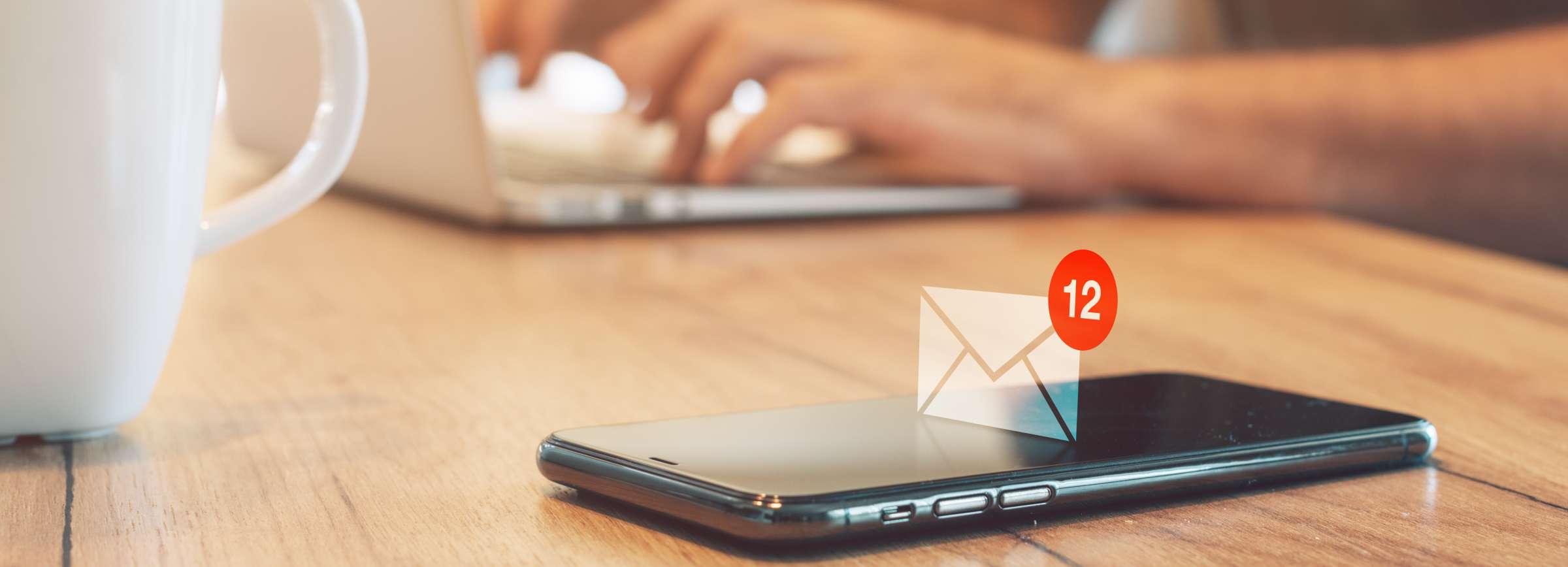 close up image of a phone laying on a table with a student typing on a laptop in the background. Floating above the phone is an email notification symbol.