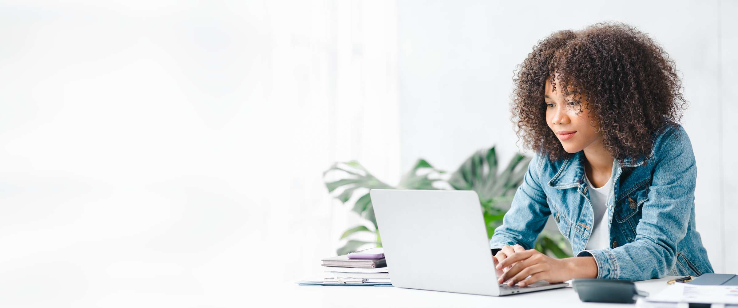 College student (Black, female, hair down and curly) sitting at a desk in a bright white room, looking down at her laptop