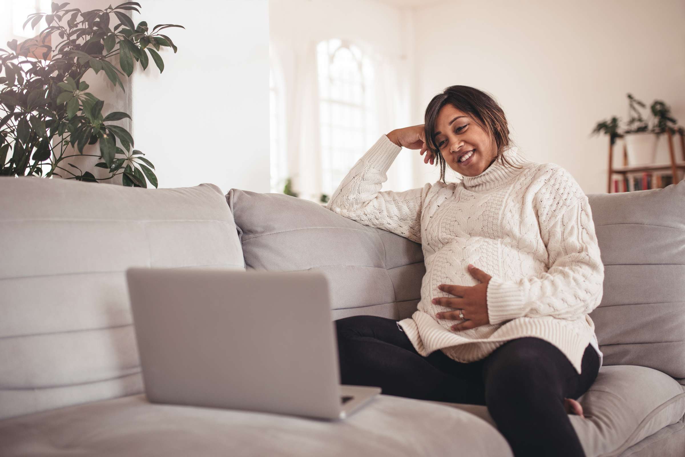 A pregnant woman sitting comfortably on a light gray couch, smiling while looking at a laptop in front of her.