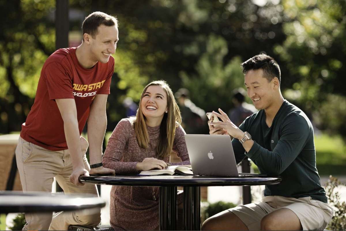 Three students around a table outside, smiling and laughing as one reads a textbook and another looks at his phone and computer