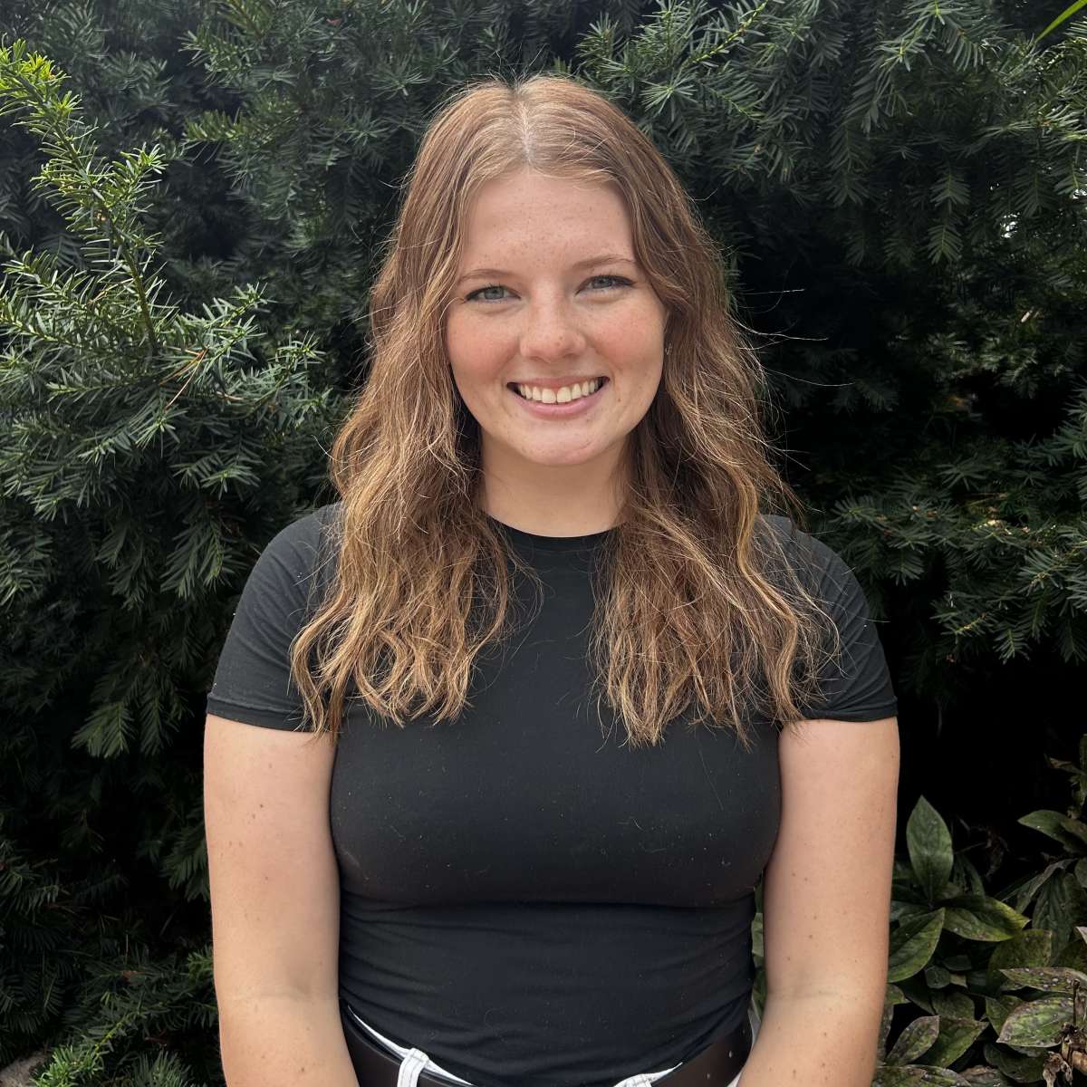 A picture of Abby, a young adult woman, with blond hair, wearing a black shift in front of greenery. 