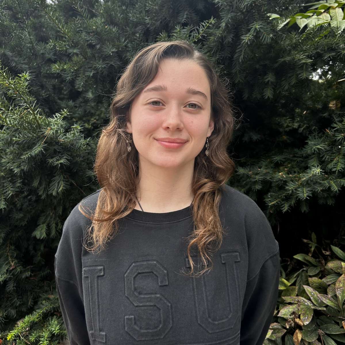 A picture of Carolyn, a young adult woman, with brown hair, wearing a black Iowa State University sweatshirt in front of greenery. 