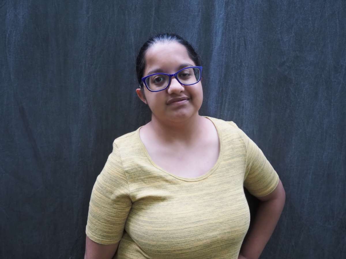 A picture of a young woman wearing a yellow top and glasses, smiling and standing in front of a black background sheet. 