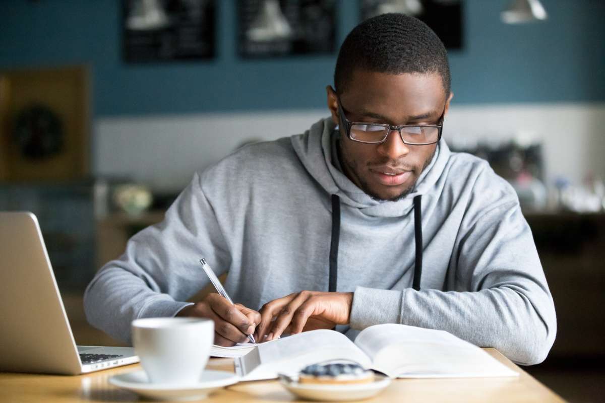 Photo of student at a coffee shop writing in a textbook with a laptop on their left wearing glasses and a sweatshirt