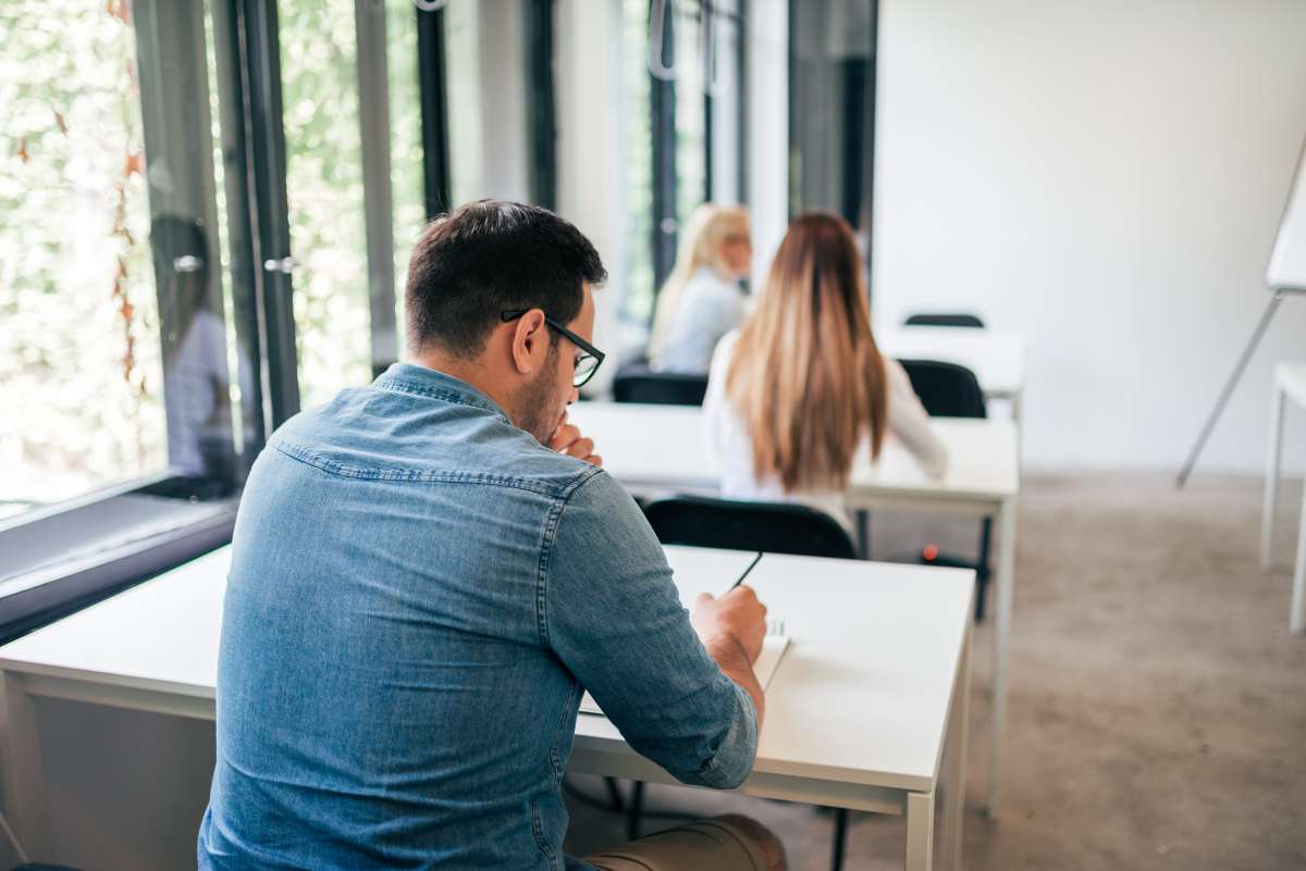 Photo taken from behind a male student, sitting at a desk taking an exam with other students in further in the background doing the same