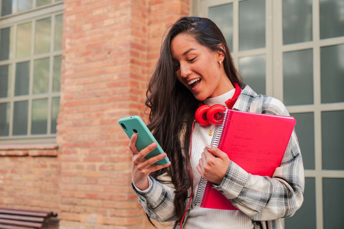 Female student with dark hair and bright read headphones holding a folder, smiling down at her blue phone outside of a brick building