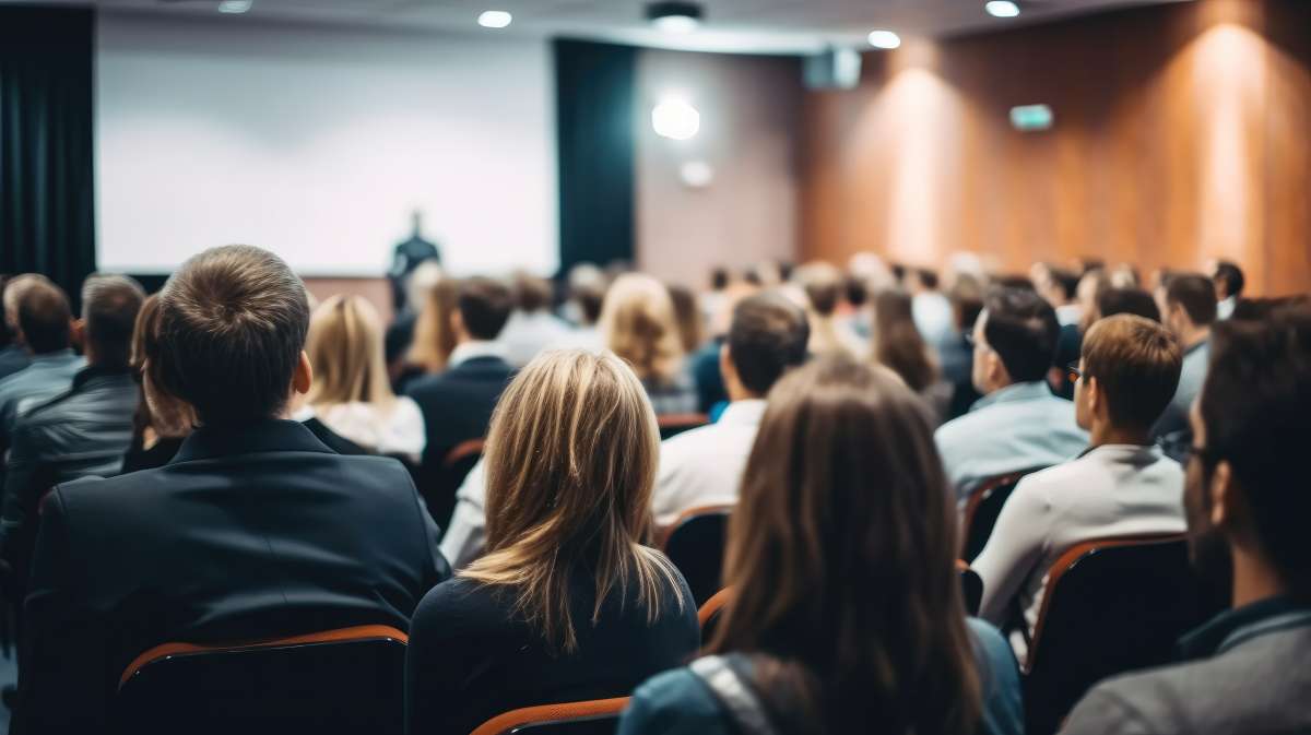 View from behind of people in audience at a lecture hall, an instructor is seen in the distance at the front speaking to the class