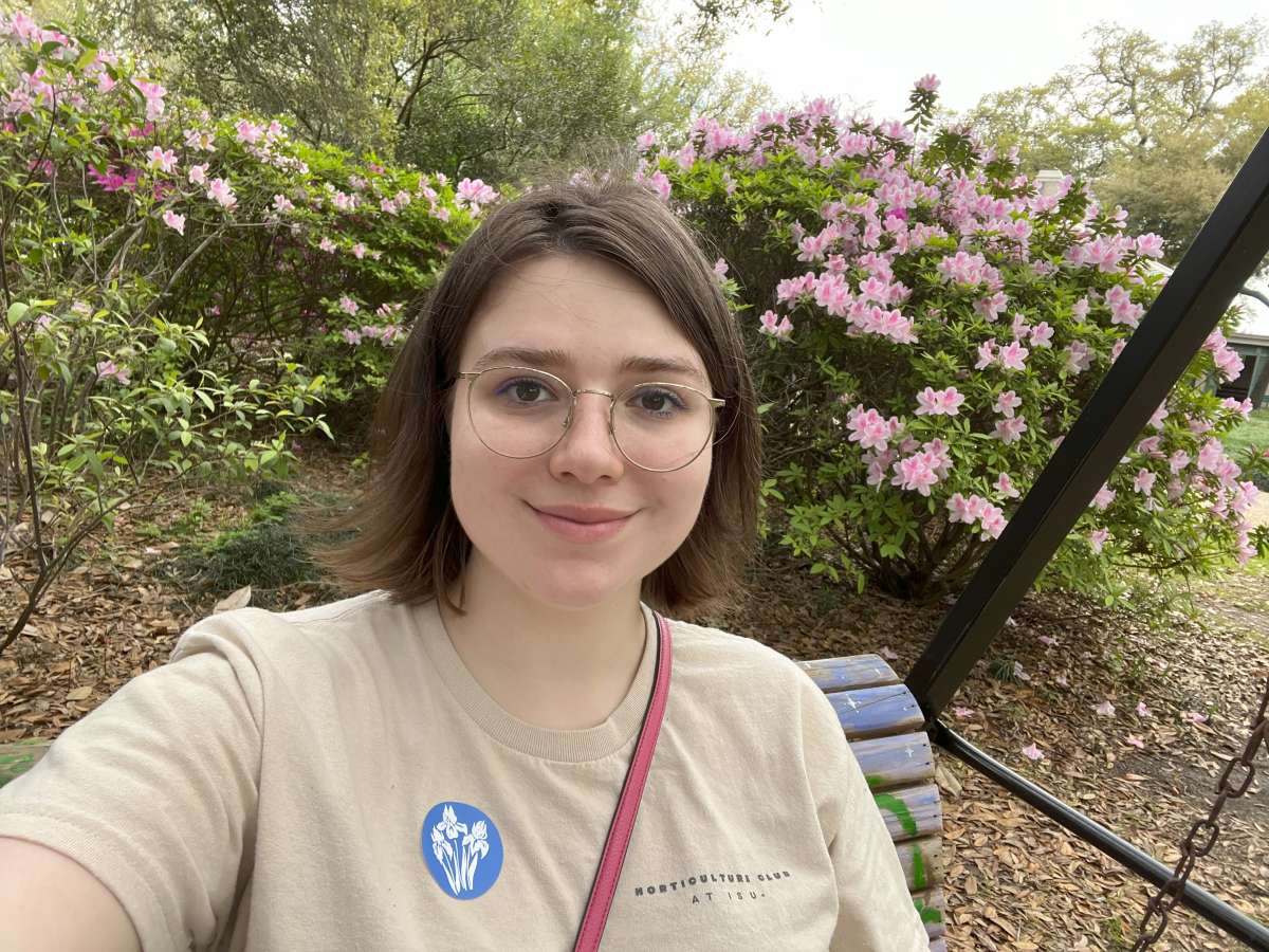 A young woman with short brown hair and round glasses is sitting on a swing bench in a garden, smiling at the camera. She is wearing a beige T-shirt with a blue sticker and has a pink strap across her shoulder. Behind her are blooming bushes with pink flowers and green foliage, and the ground is covered in fallen leaves. 