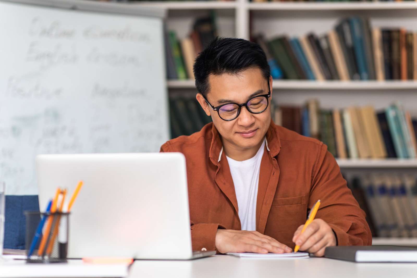 Man with black hair and glasses sitting in front of a laptop, looking down while writing on paper