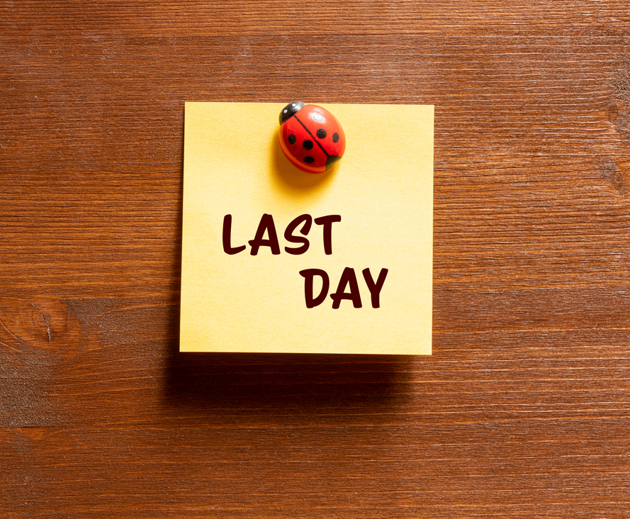 Yellow sticky note stuck on a wooden background that says, 'Last Day' and is pinned with a ladybug pin